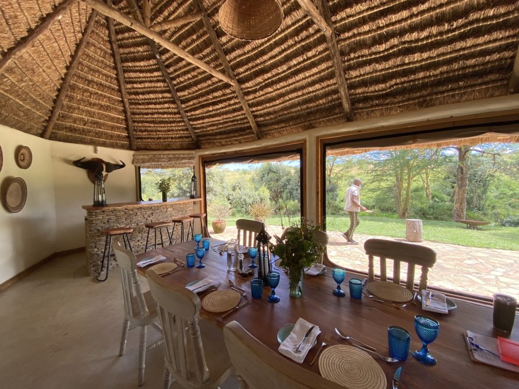 Long wooden dining table set with blue glassware beneath a thatched roof, beside a stone bar at El Karama Lodge.