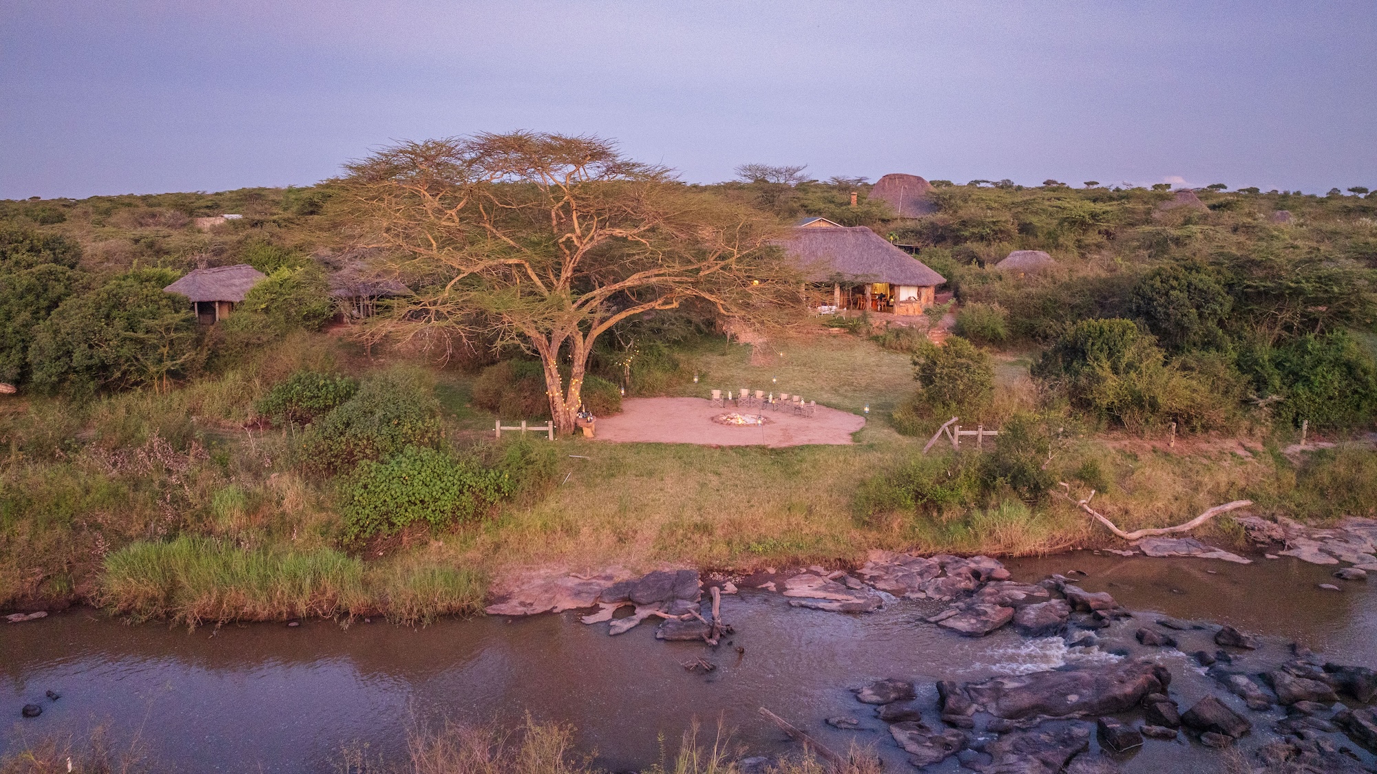 Aerial view of El Karama Lodge at dusk, with a fire pit and chairs beneath a fever tree above the rocky Ewaso Nyiro River.
