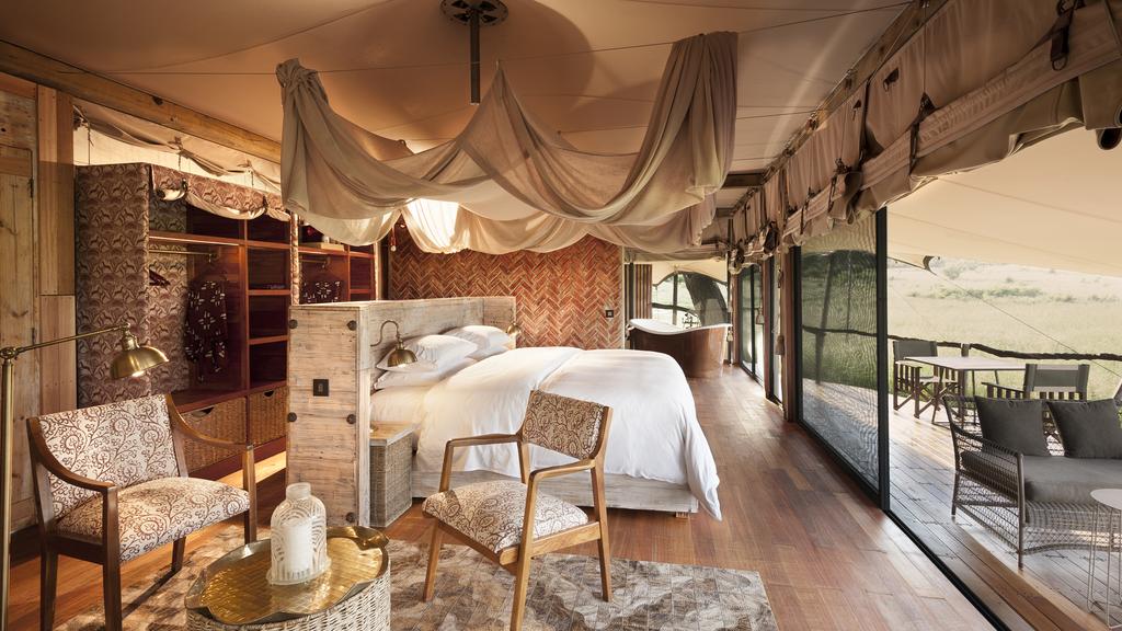 A spacious, elegantly appointed guest tent at Somalisa Bush Camp, featuring a king bed draped with gathered canvas canopy, herringbone brick feature wall, open wardrobe with patterned wallpaper, polished hardwood floors, and floor-to-ceiling mesh walls opening onto a private deck overlooking the Hwange bush, Zimbabwe.