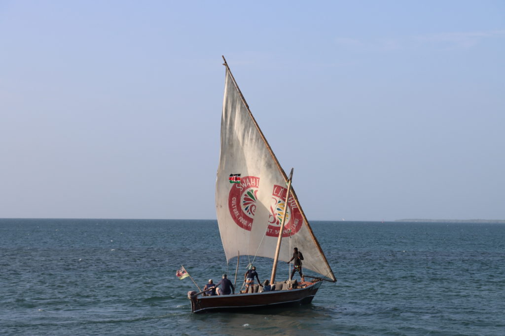 Traditional wooden dhow under a white lateen sail flying the Kenyan flag, with crew and guests aboard in the Lamu Archipelago.