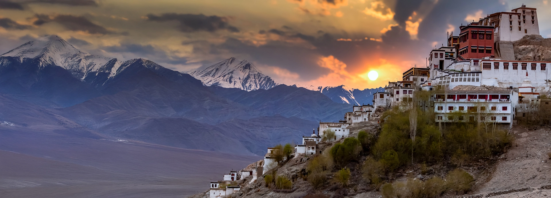 A Himalayan monastery complex perched on a hillside in Ladakh at sunset, overlooking a stark mountain landscape bathed in golden light.