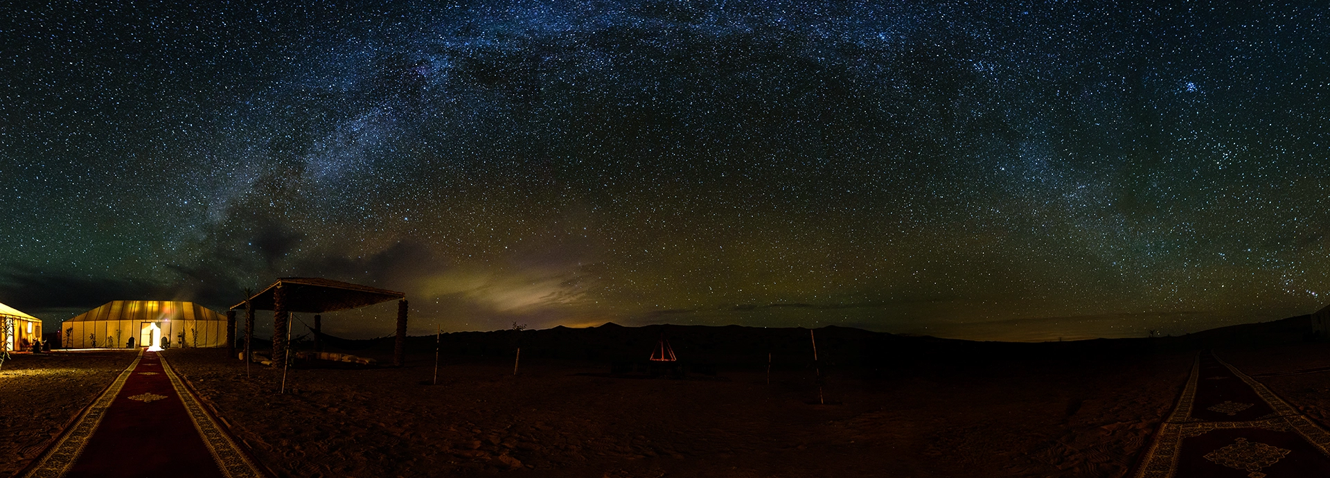 The Milky Way galaxy stretching across the night sky above glowing tents in the Moroccan Sahara desert.