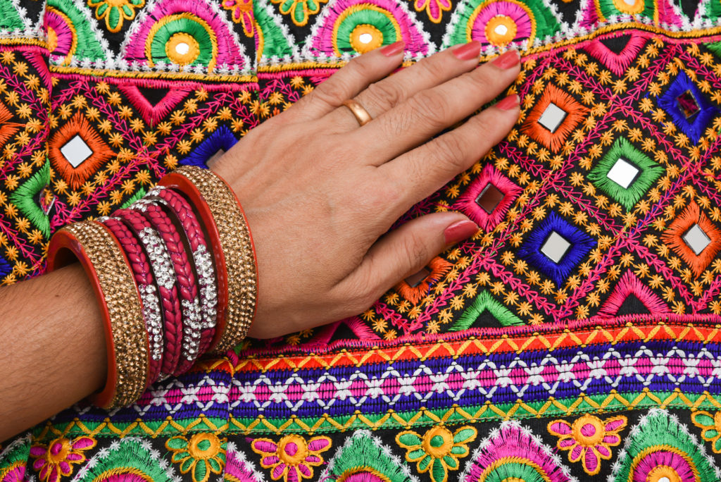Close-up of a hand wearing bangles resting on vividly embroidered, mirror-work fabric