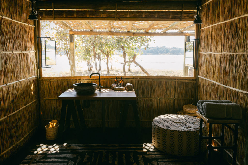 An open-air bathroom at Kakuli, its reed walls framing a wood-topped vanity and hand-thrown basin, with the Luangwa River visible through the trees beyond.