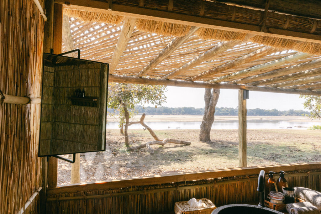 Morning light catches a round mirror and washstand in a reed-walled bathroom, the river and floodplain stretching out beyond the open frame.