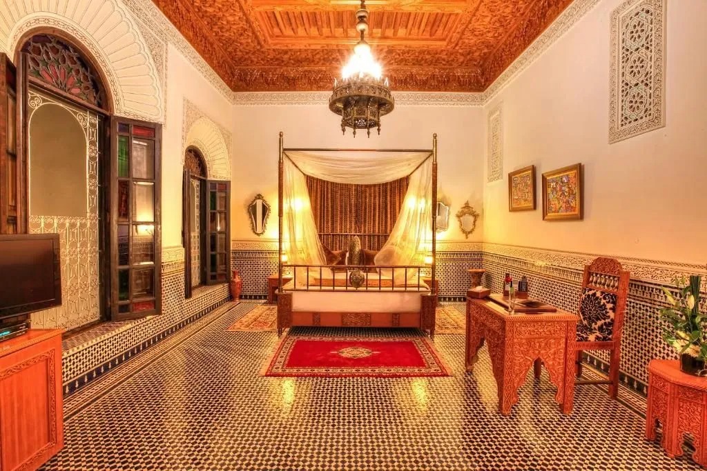 An Ambassador Suite with a carved four-poster bed draped in sheer curtains beneath an intricately carved cedar ceiling, tiled floors and a red Moroccan rug underfoot.