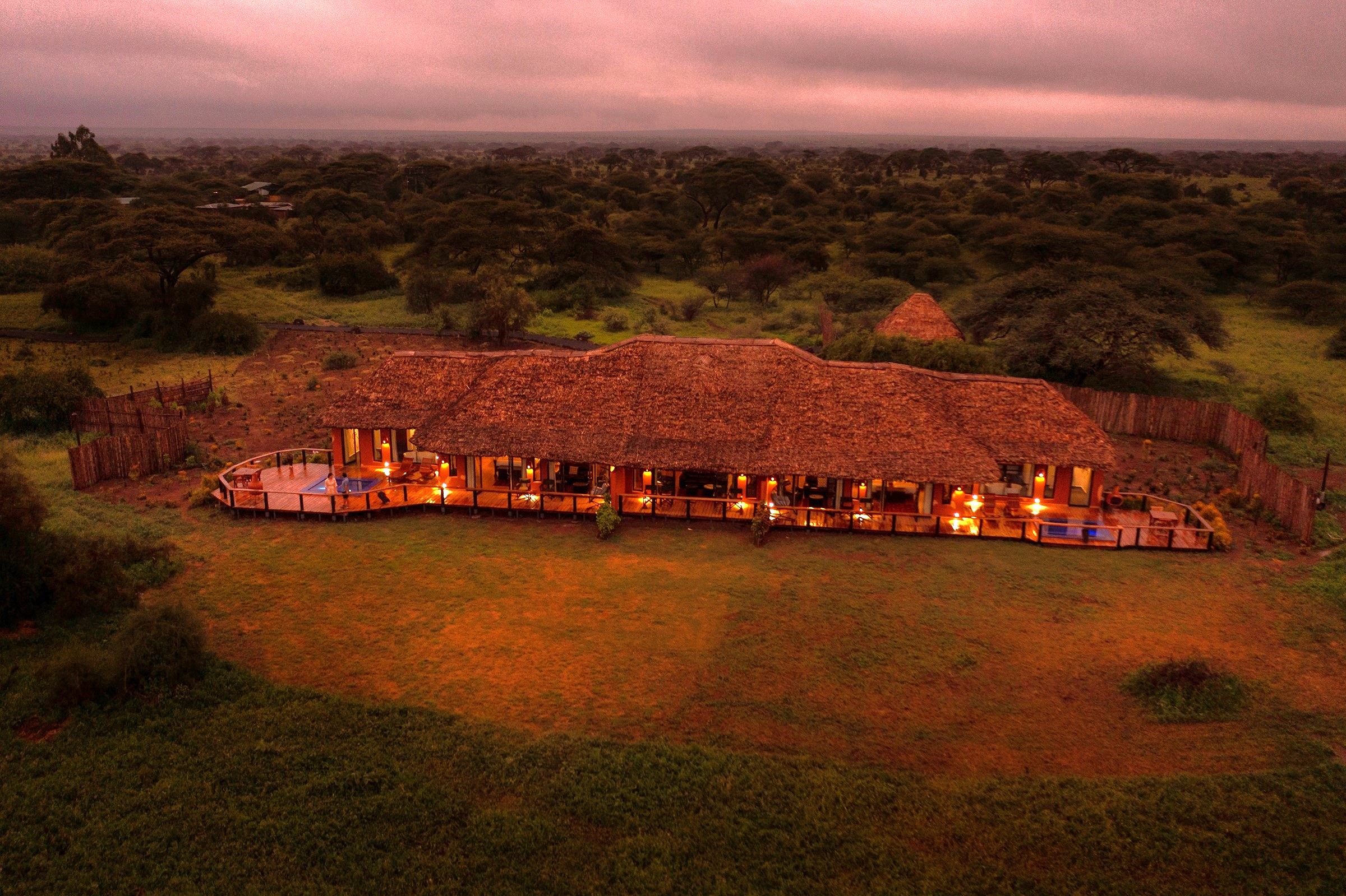 Aerial view of a thatched Tawi Lodge suite at sunrise, its lit deck and plunge pools facing acacia woodland in Kenya's Amboseli region.