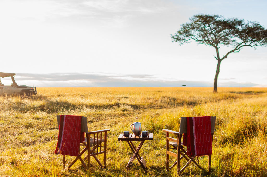 Two canvas director's chairs draped with red Maasai blankets face out over golden grassland at sunrise, a small camp table holding a coffee pot and mugs set between them, with a safari vehicle and lone acacia tree in the distance.