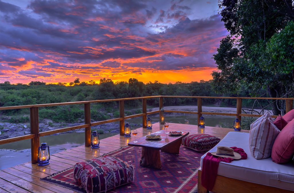 A riverside deck set with cushions, lit lanterns and glasses of rosé wine, framed by trees, glows beneath a dramatic orange and purple sunset over the Talek River.