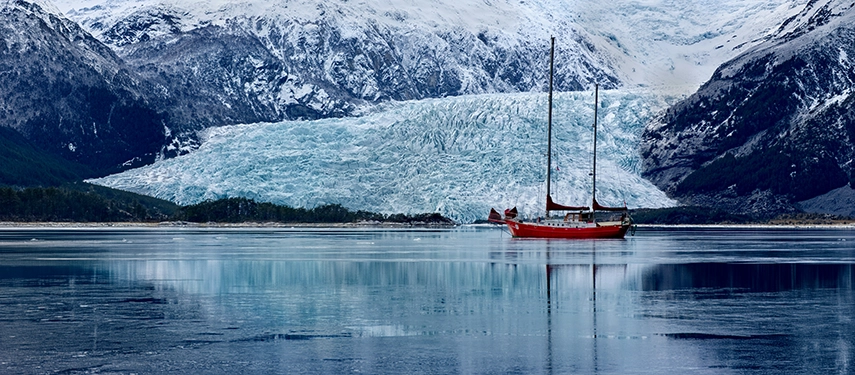 A red expedition yacht anchored before a vast Patagonian glacier, exploring the remote fjords of southern Chile.