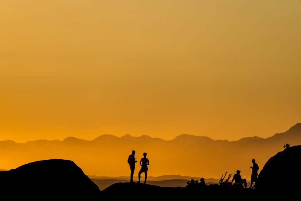 Guests gather at sundowner hour on the boulders above Mowani Mountain Camp, silhouetted against a molten Damaraland sky.