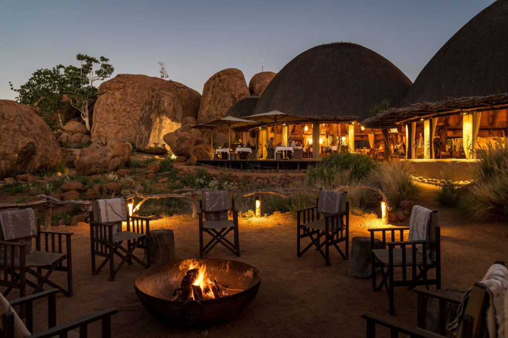 Lantern-lit dining tables glow beneath Mowani's thatched roofs at dusk, with an empty firepit and director's chairs waiting in the foreground.