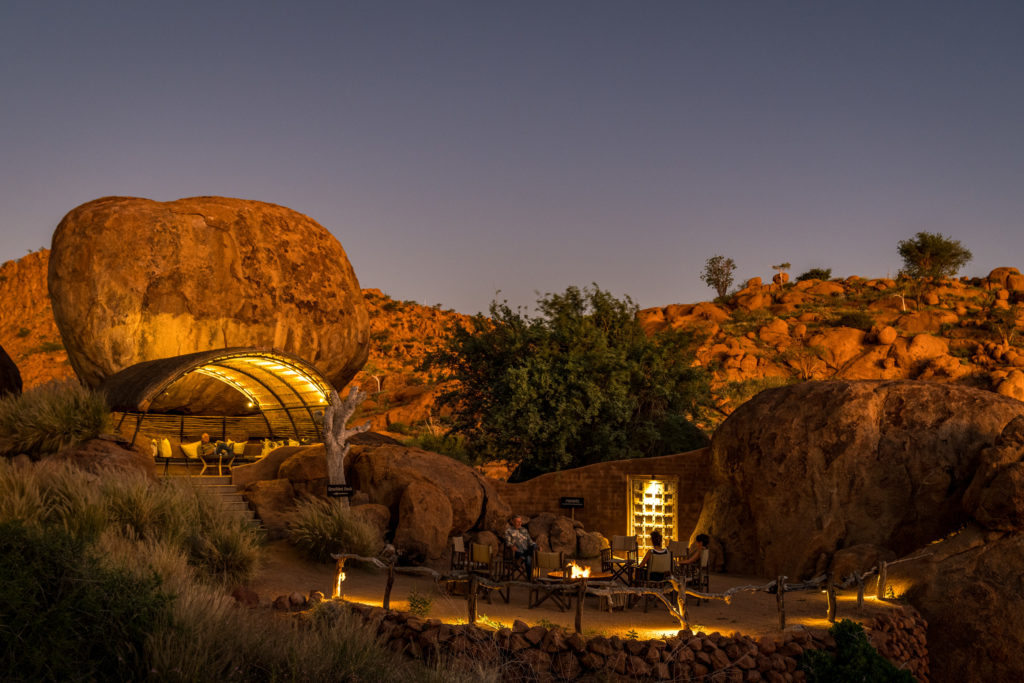 Guests relax in a boulder-shaded lounge at Mowani as evening falls, with a firepit and circle of chairs set out below.