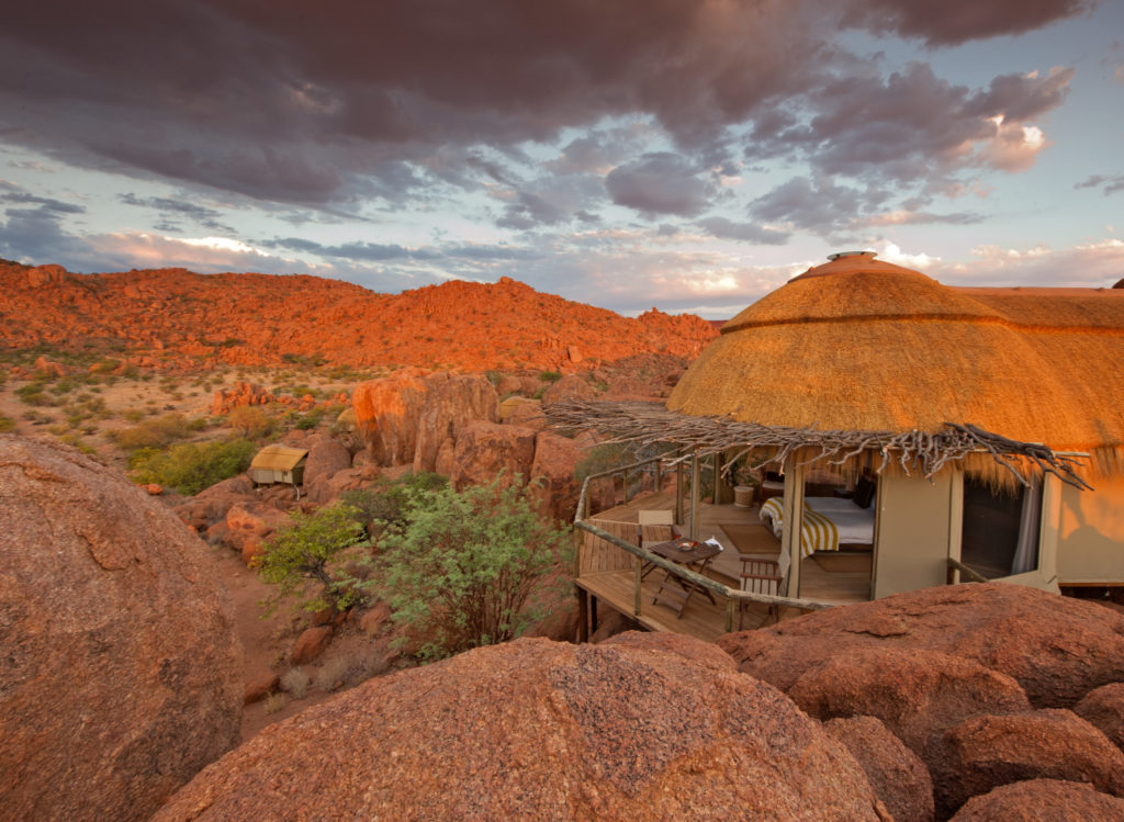 A Mowani suite's deck and bed catch the fading light, framed by dramatic dark clouds over the surrounding boulder-strewn hills.