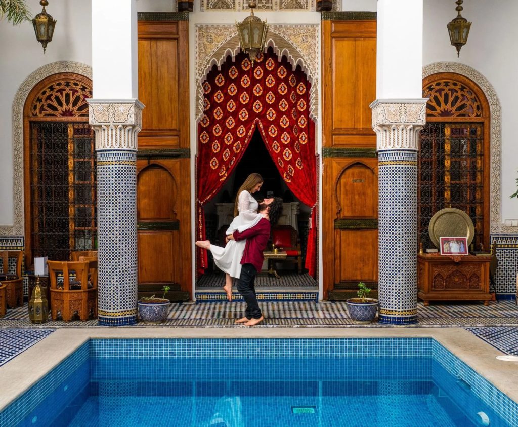 A couple embraces beside the courtyard pool of Riad El Amine Fès, framed by zellige-clad columns and a gold curtained doorway in the heart of the Fès medina.