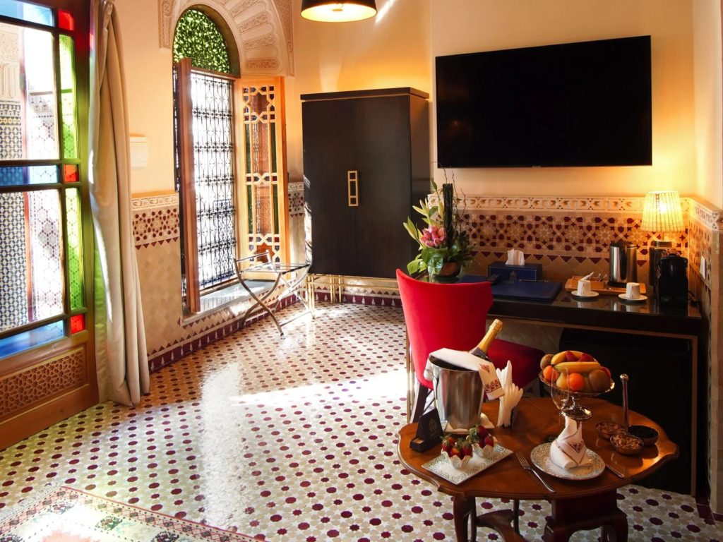 A suite corner with a dark wooden armoire, stained-glass door and a round table laid with champagne and fresh fruit.