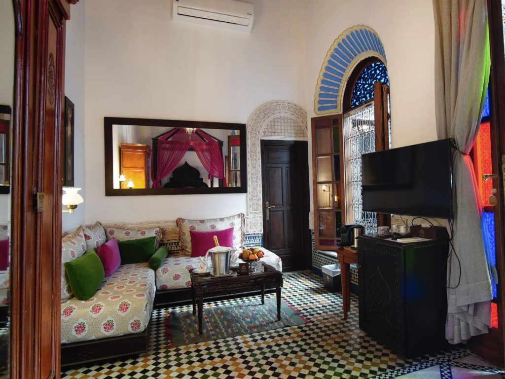 A sitting area with a floral banquette in pink and green tones, a gilt-framed mirror reflecting the canopy bed beyond, and a fan-shaped stained-glass window.