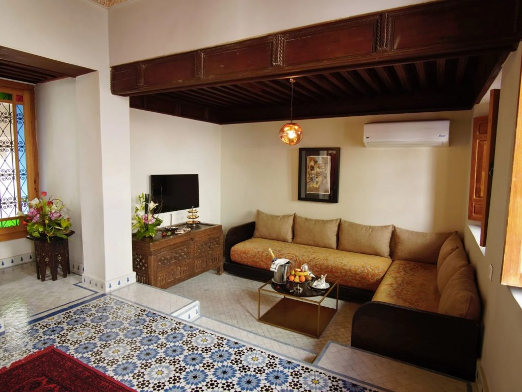A living space beneath a carved wooden ceiling beam, with a mustard-toned sofa, patterned tilework and traditional artwork on the walls.