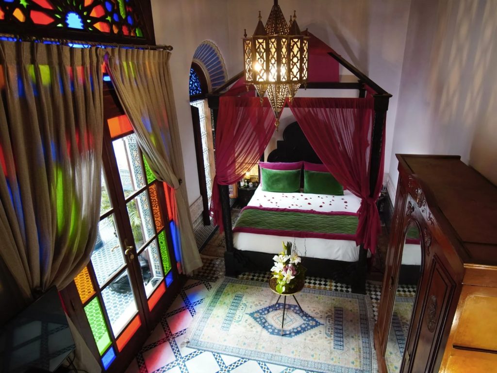 A canopy bed dressed in burgundy drapes and scattered rose petals, lit by a brass lantern chandelier and framed by stained-glass windows.