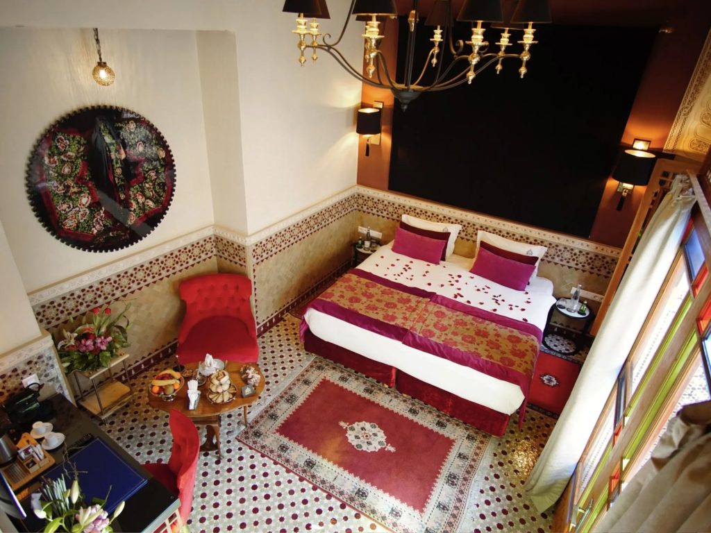 An aerial view of a suite bedroom, with burgundy and gold bedding scattered with rose petals and a tufted red armchair beside intricate mosaic flooring.
