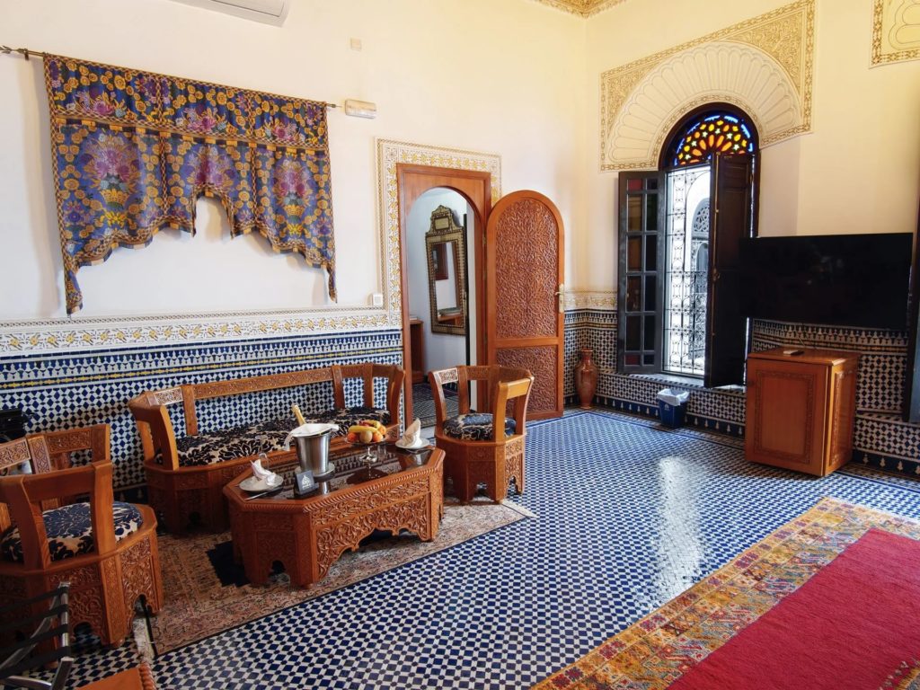 A living room with an embroidered curtain valance, carved wooden doors and a stained-glass arched window overlooking the medina.
