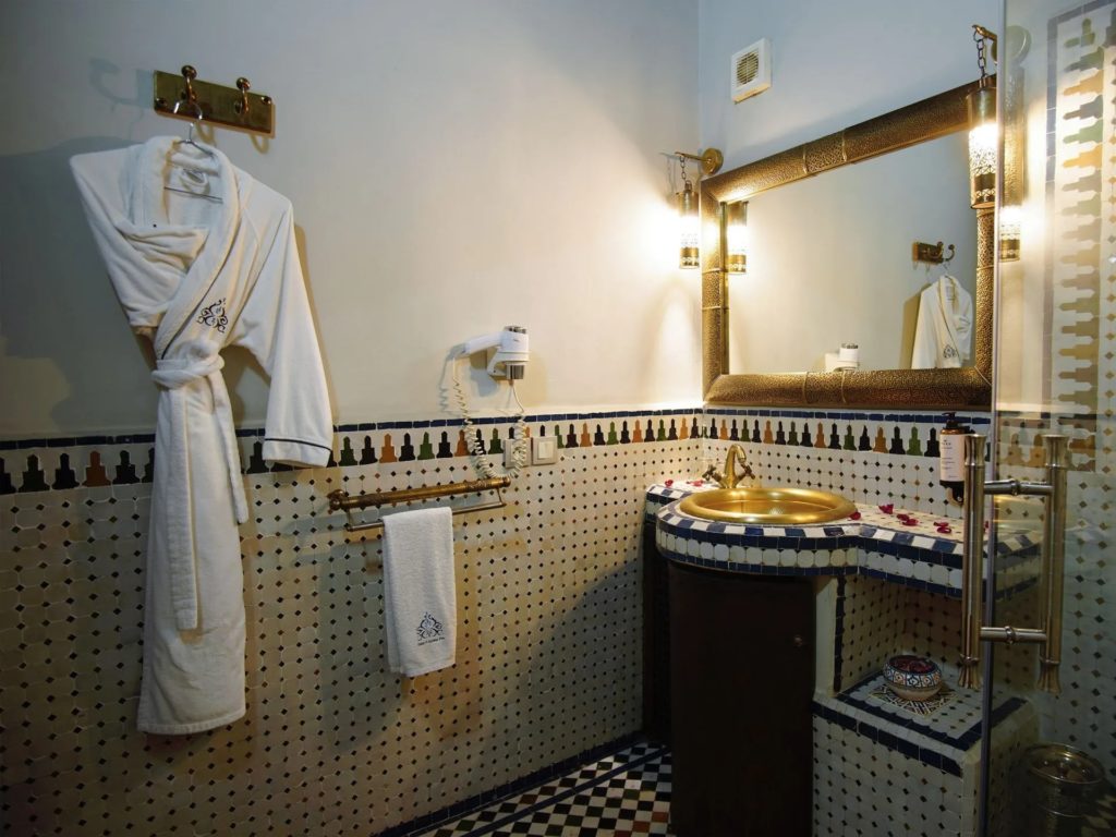 An en-suite bathroom at Riad El Amine Fès, with a hand-hammered brass basin, mosaic tilework and a plush robe hung ready for guests.