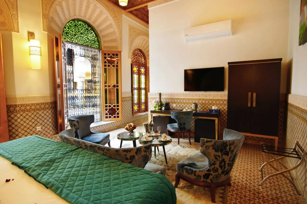 A bedroom dressed in deep green linens, with blue velvet armchairs, stained-glass doors and carved plaster archways.