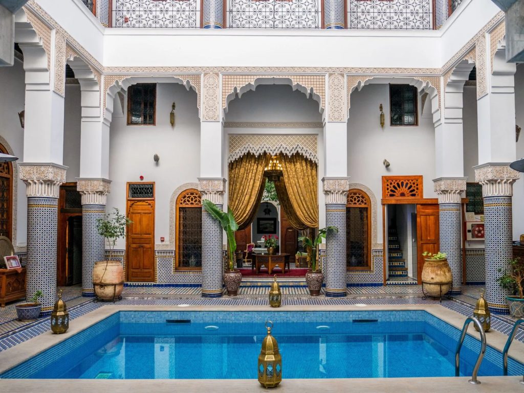 The courtyard pool of Riad El Amine Fès, framed by zellige columns, potted palms and golden drapery beneath a carved plaster archway.