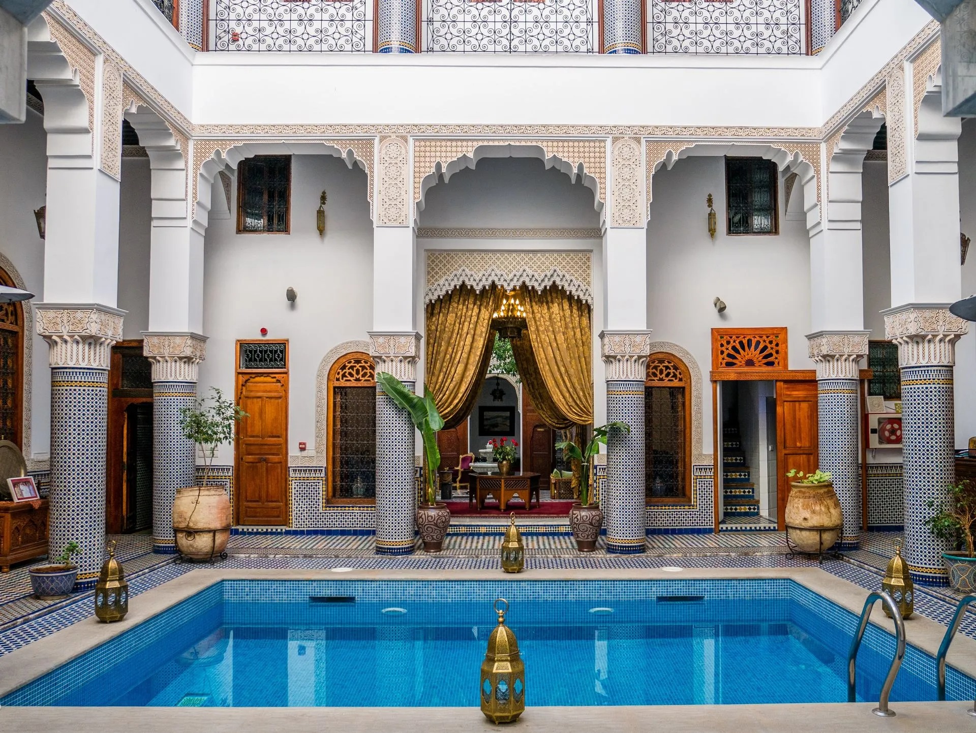 The courtyard pool of Riad El Amine Fès, framed by zellige columns, potted palms and golden drapery beneath a carved plaster archway.