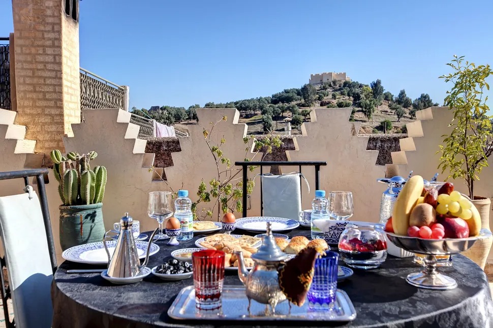 A rooftop breakfast spread of pastries, tea and fresh fruit, overlooking the hills and historic fortifications surrounding Fès.