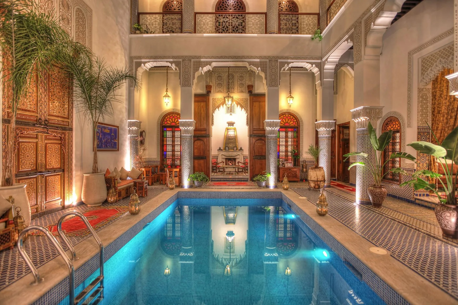 The illuminated courtyard pool of Riad El Amine Fès at night, framed by carved columns and a glowing fireside lounge beyond.