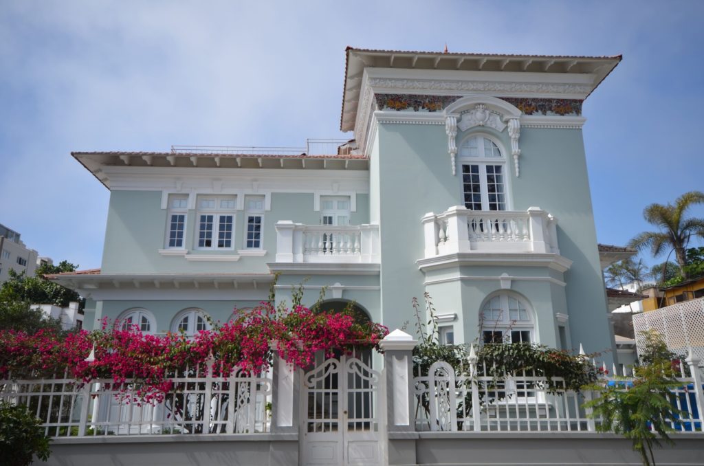 Pastel-blue colonial mansion facade with white balustraded balconies and bougainvillea climbing a wrought-iron gate