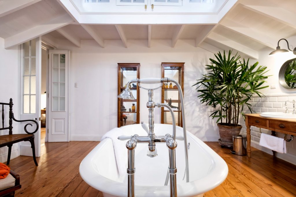 Freestanding white bathtub with chrome clawfoot taps beneath a skylight, framed by potted palms and antique display cabinets