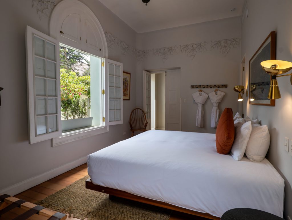 Bright bedroom corner with an open shuttered window, terracotta cushion and folded white robes hanging on the wall