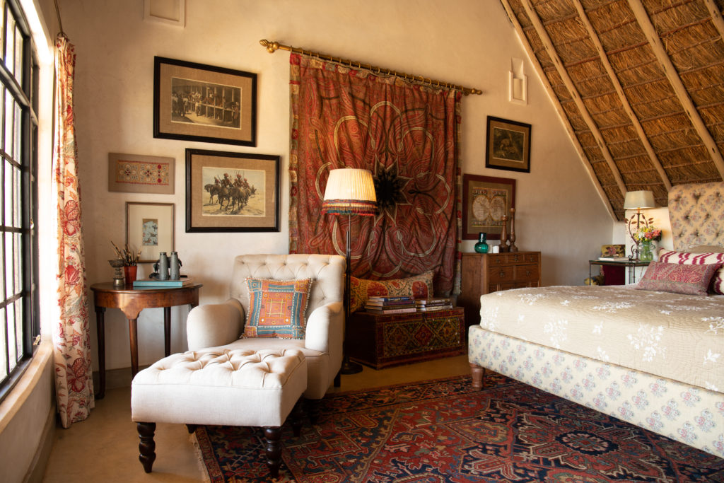Bedroom corner at Olepangi Farm with a tufted armchair and footstool, framed prints, a paisley wall hanging and a Persian rug under thatch.