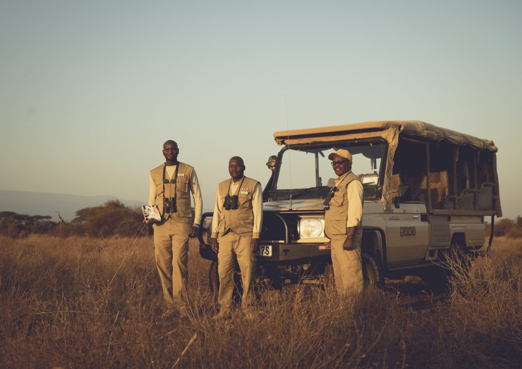 Three guides in khaki with binoculars stand beside an open-sided four-wheel drive game drive vehicle in low golden light.