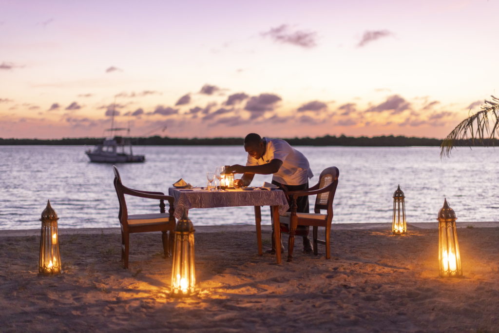 Staff member lights a lantern on a beach dinner table for two, ringed by brass lanterns on the sand at dusk, Manda Bay, Kenya.