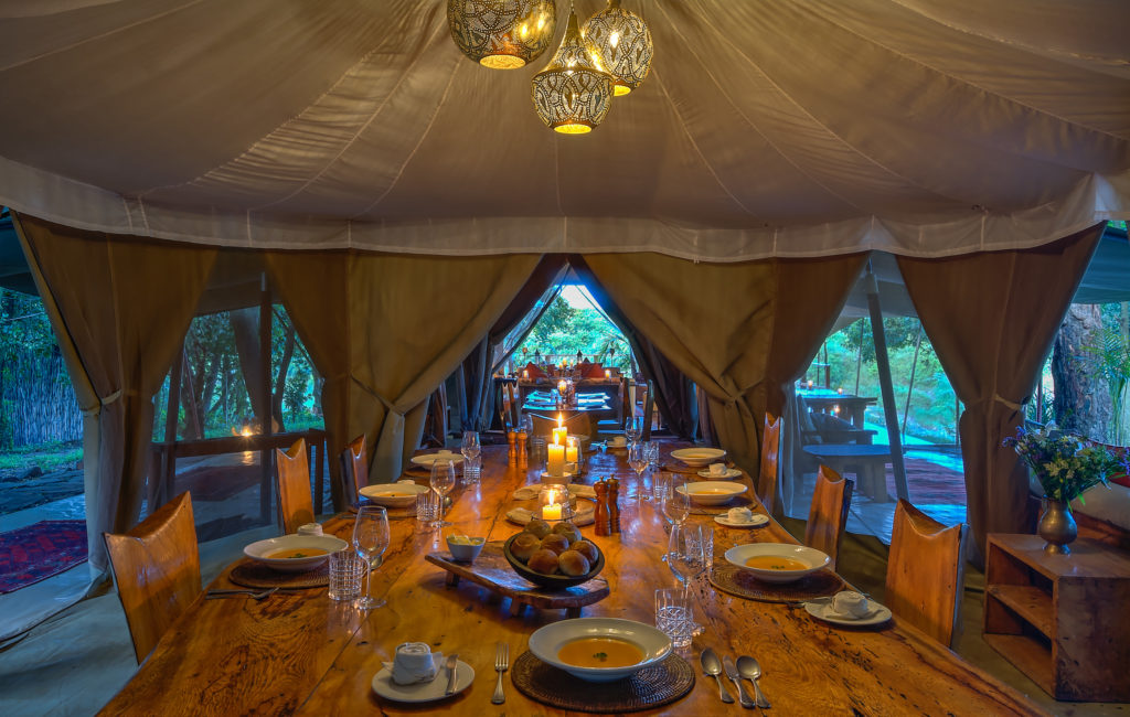 Inside Naibor's candlelit dining tent, a long wooden table is laid with bowls of soup, bread and glassware beneath hanging lantern-style lights, with further dining areas visible through the open canvas doors.
