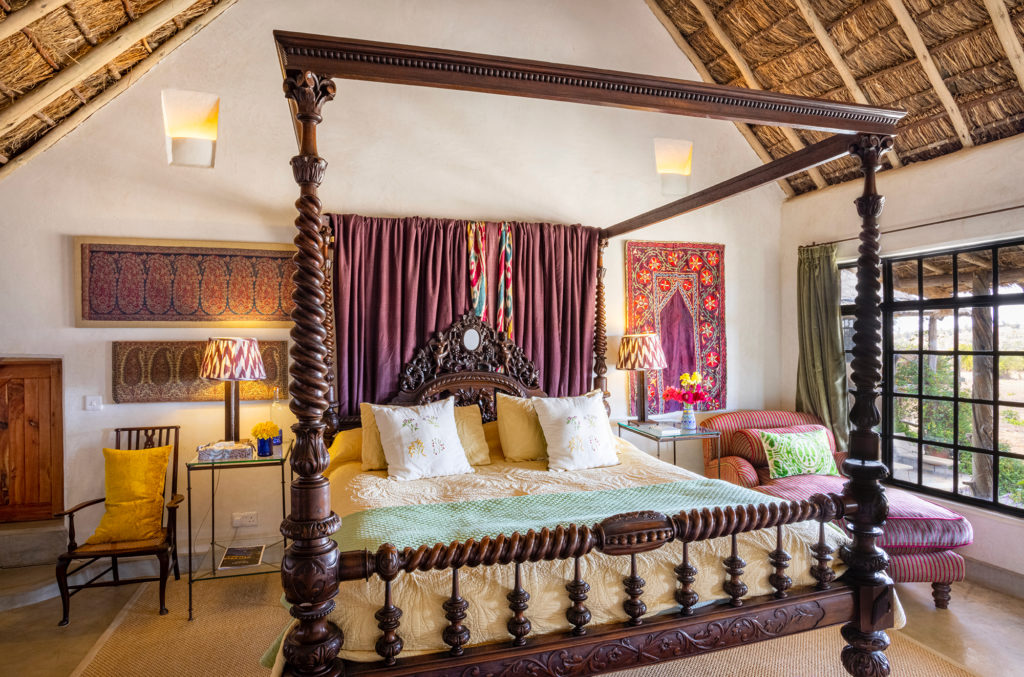 East Wing bedroom with a carved four-poster bed, suzani wall hanging, ikat lampshades and a thatched roof at Olepangi Farm, Laikipia.