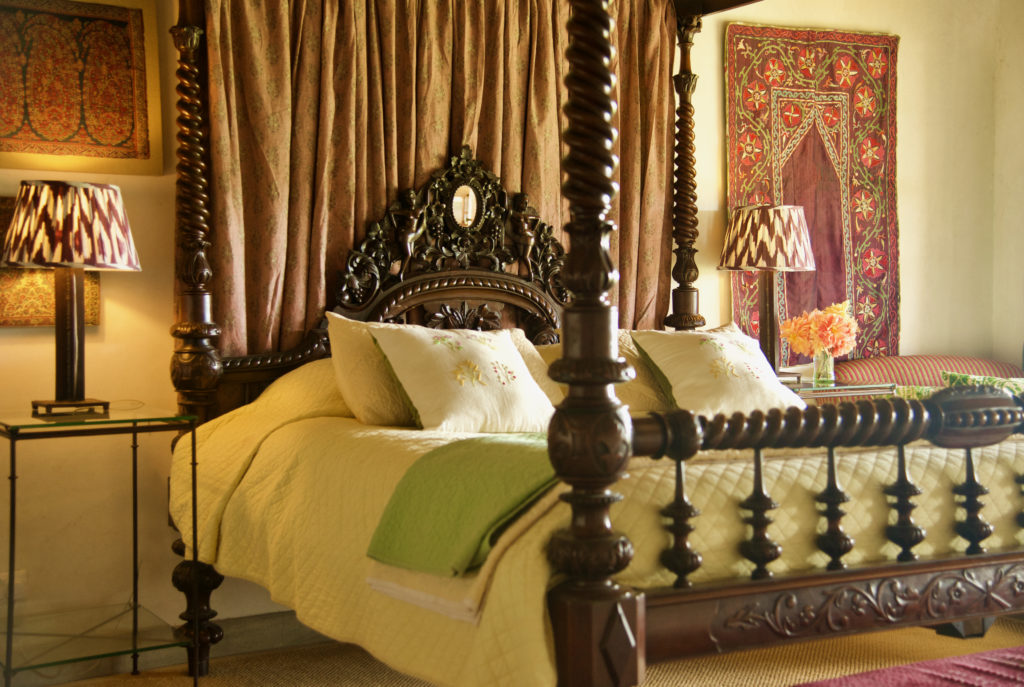Close view of a carved barley-twist four-poster bed with embroidered cushions, a green throw and orange roses in warm lamplight.