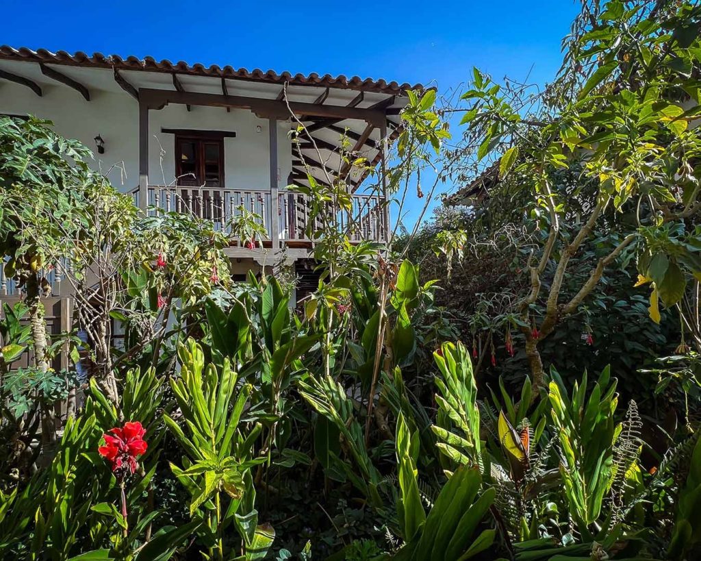 Tiled hotel building glimpsed through lush garden foliage and flowering shrubs beneath a bright blue sky