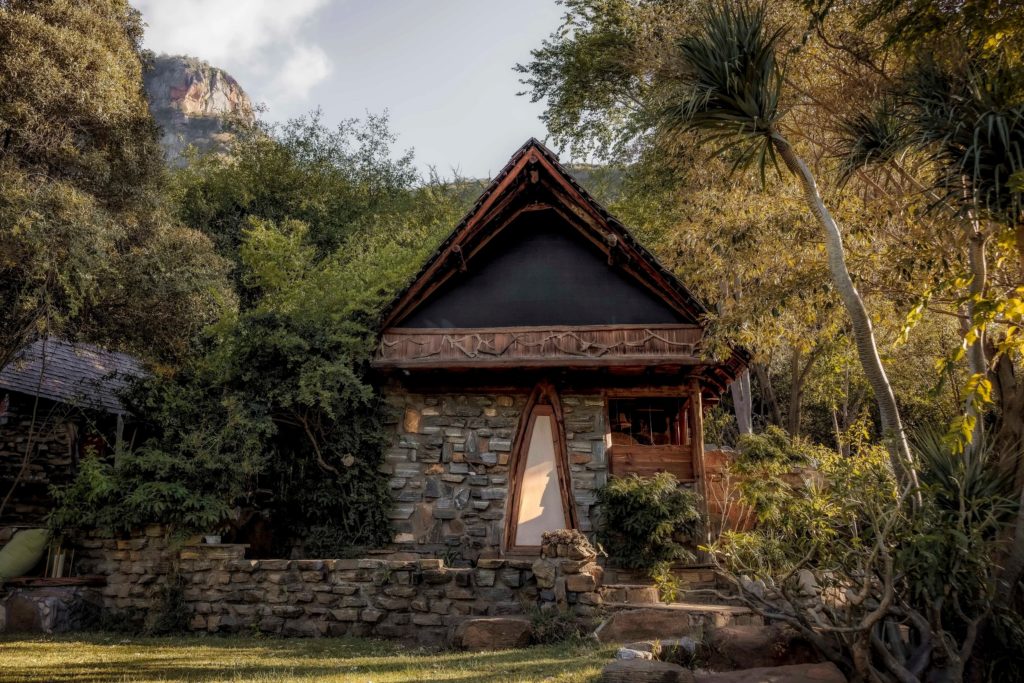 Back of the family villa at Desert Rose, with dry-stone walls, a steep timber gable and a rock cliff rising behind in afternoon light.