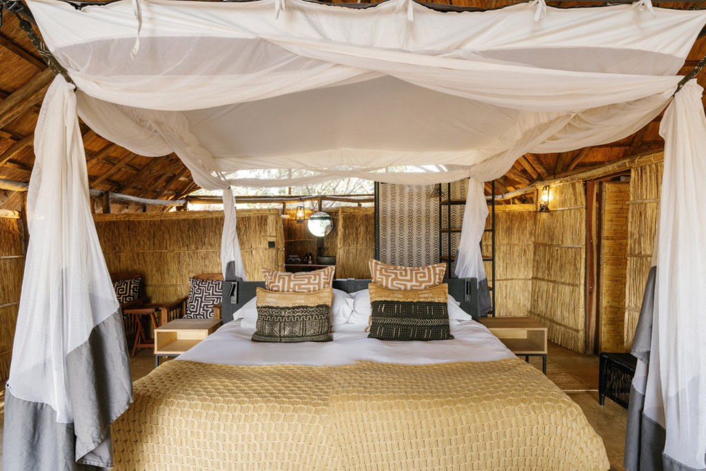 A four-poster bed dressed in mud-cloth cushions and pale linen sits beneath billowing white netting, reed walls glowing warm in the light.