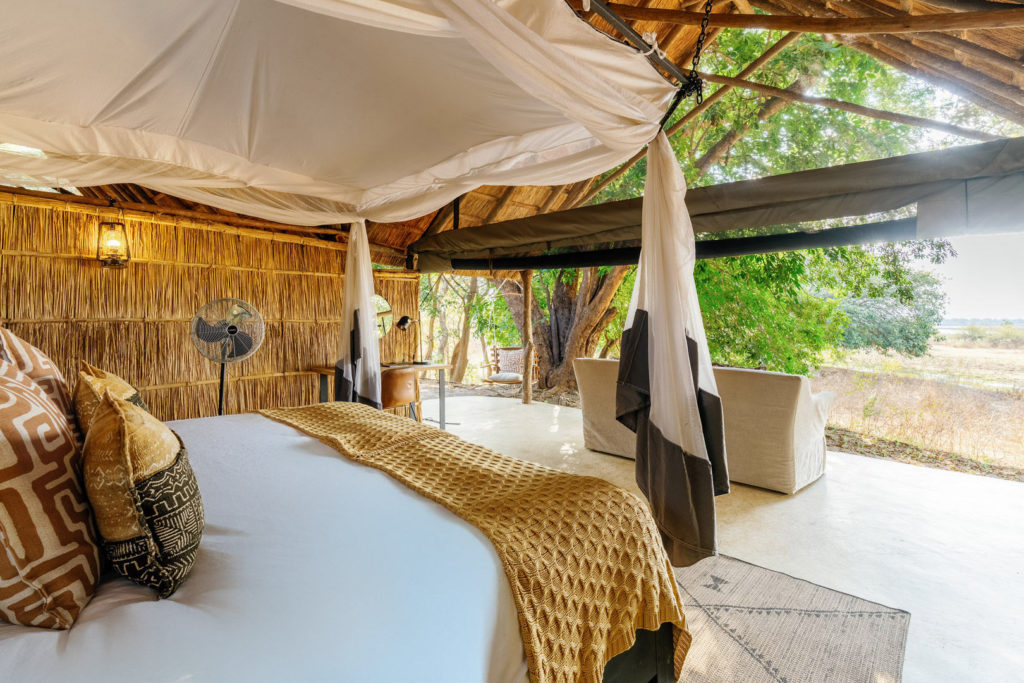 Draped mosquito netting frames a comfortable bed that opens onto a private lounge area, greenery visible just beyond the canvas edge.