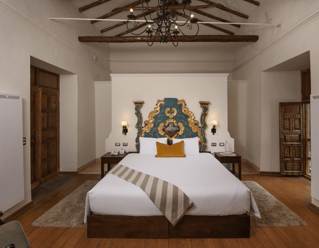 Suite with an ornate gilded and turquoise headboard beneath dark timber ceiling beams and a wrought-iron chandelier