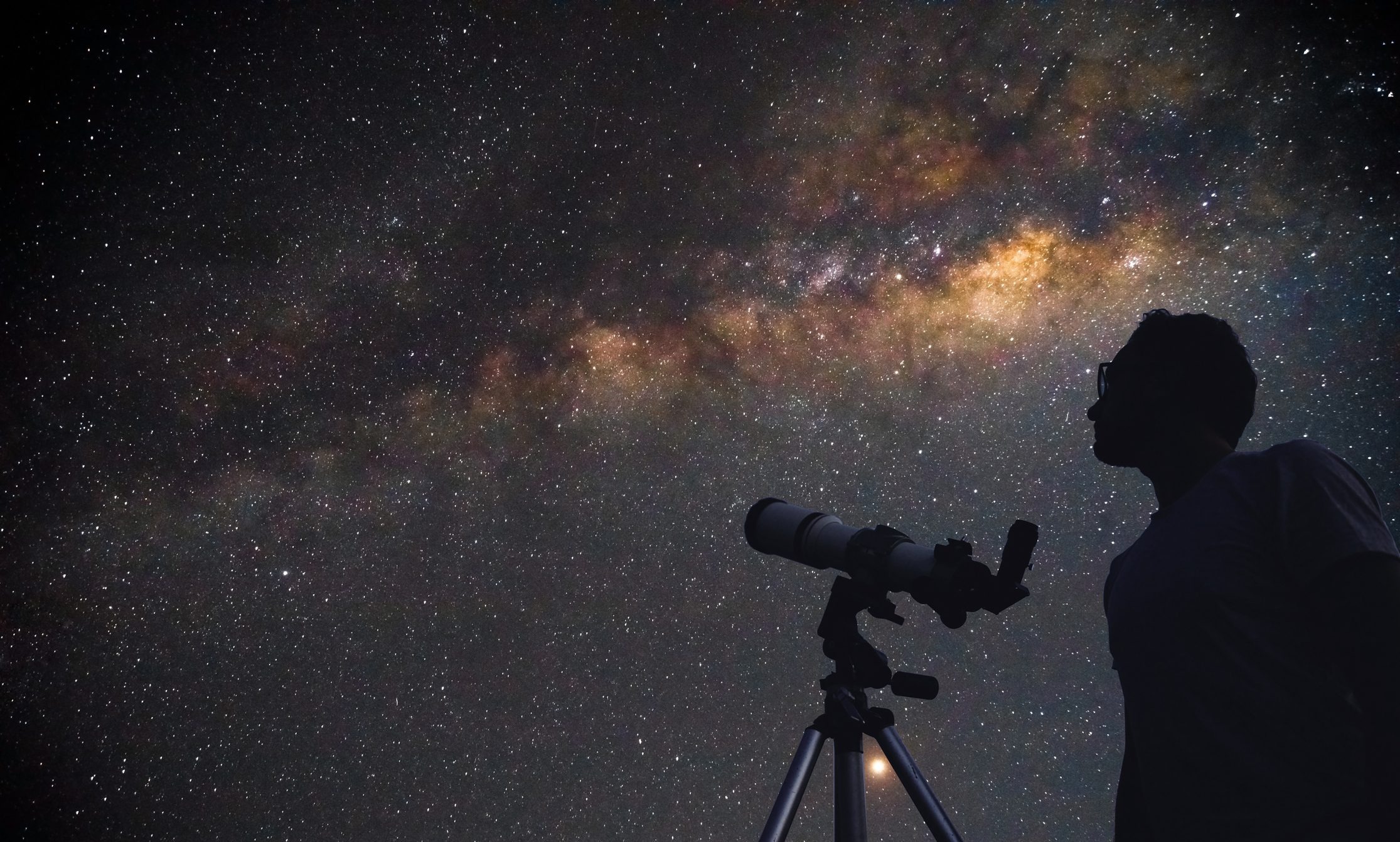 A stargazer sets up a telescope beneath the Milky Way, the desert sky thick with stars.