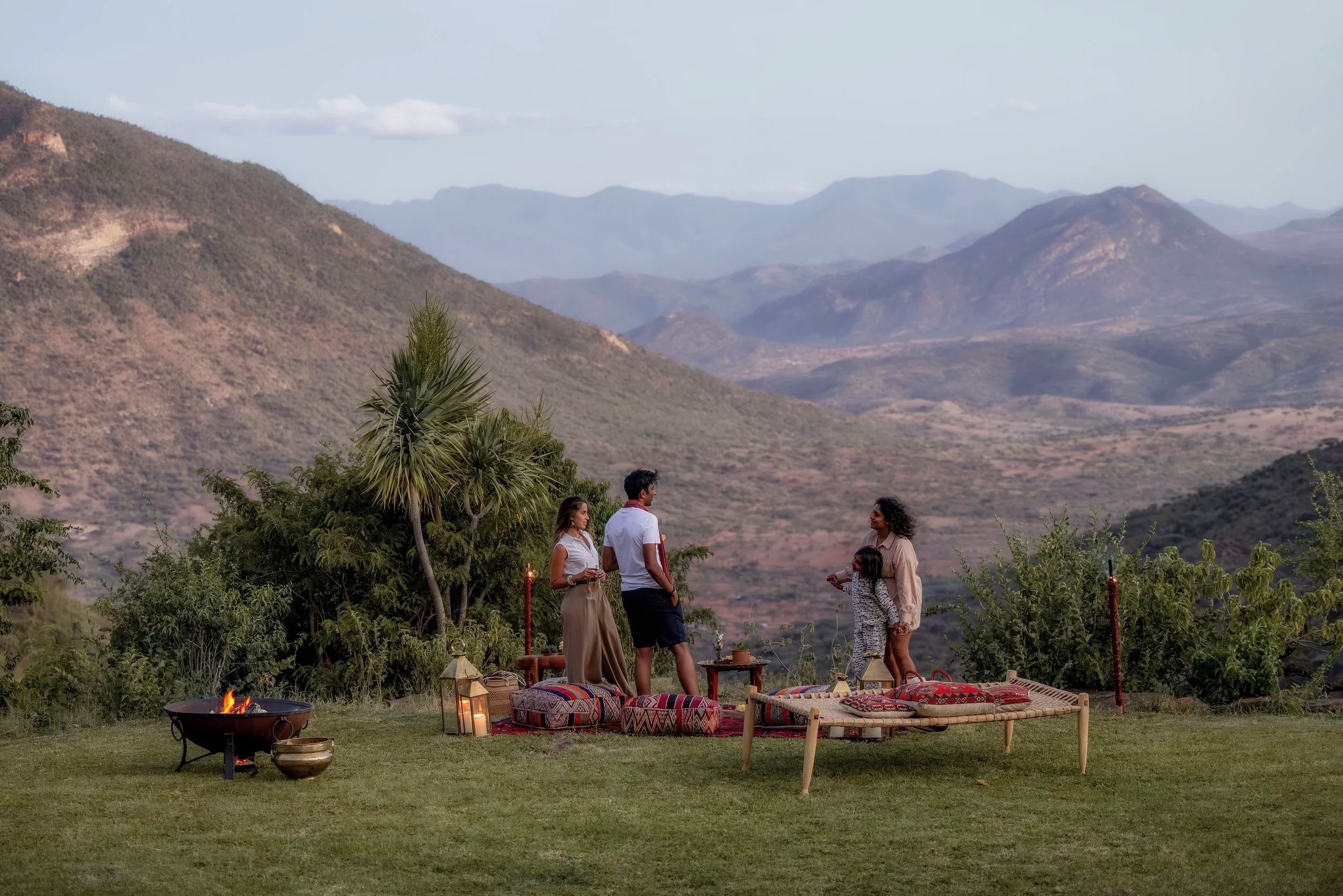 A family gathers for a sundowner beside a fire pit, floor cushions and a woven daybed above a wide valley at Desert Rose.