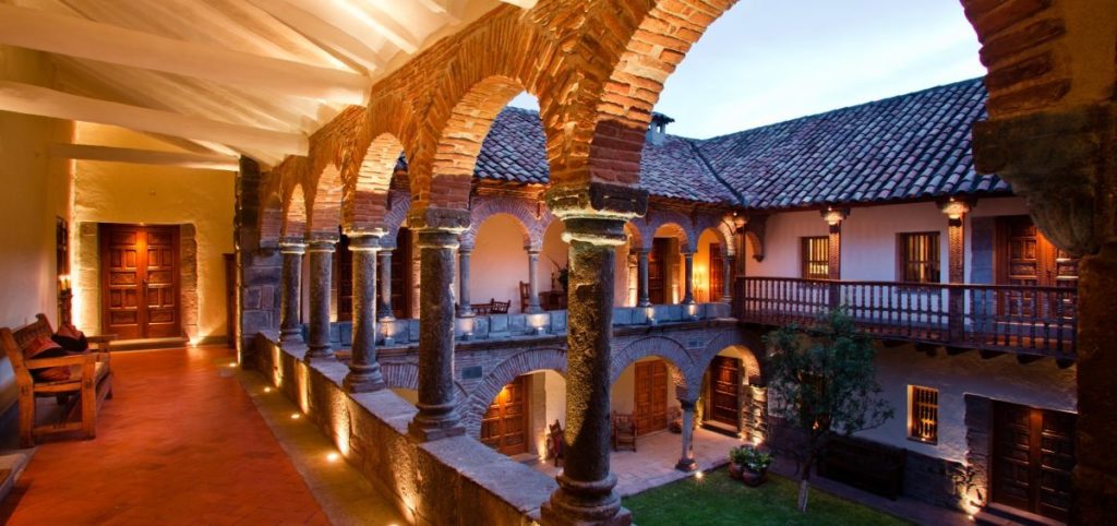 Illuminated stone cloister at dusk with arched colonnades, a carved wooden balcony and terracotta roof tiles