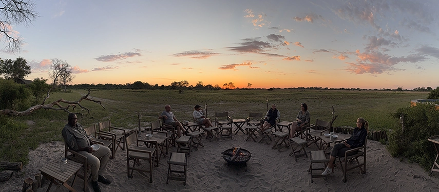 Guests gather around a firepit at sunset, sharing drinks and conversation during a relaxed safari sundowner set against open savanna and fading evening light.