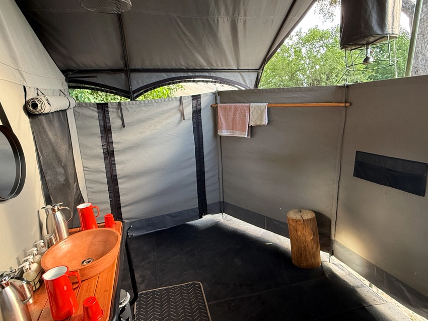 An open-air safari bathroom features a canvas-walled shower and hand-carved basin, blending simplicity with thoughtful comfort in the African bush. 